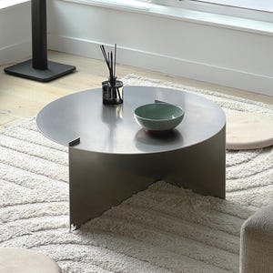 May include: A modern, semi-circular metal coffee table with a matte finish. A green and red bowl and a black diffuser bottle with reeds sit on the table. The table rests on a white, textured rug.