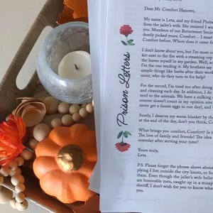 May include: A fall-themed display featuring a wooden tray with decorative elements. It includes a candle in a glass holder, an orange pumpkin, and a stack of papers with the title "Prison Letters" and handwritten text.