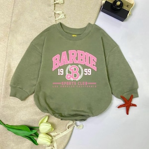 May include: Olive green baby romper with the word "BARBIE" in pink, along with "1959" and "SPORTS CLUB LOS ANGELES CALIFORNIA" in pink. The romper has long sleeves and a round neckline.