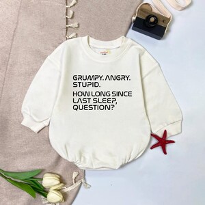 May include: A cream-colored baby onesie with long sleeves. The front of the onesie features the text: "GRUMPY. ANGRY. STUPID. HOW LONG SINCE LAST SLEEP, QUESTION?" A red starfish and a toy camera are also visible.