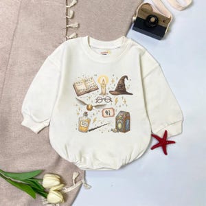 May include: Cream-colored baby bodysuit with a whimsical print featuring a book, candle, wizard hat, glasses, golden snitch, potion bottle, wand, and a number 9 3/4. The design is in shades of brown, gold, and black.