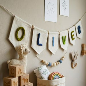 Puede incluir: Una pancarta crema con el nombre "OLIVER" en letras verdes, azules y crema cuelga de una pared. También se ven una llama de peluche, bloques de madera y un juguete de madera en forma de arcoíris.