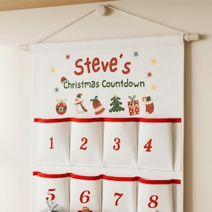 Könnte beinhalten: Ein weißer Weihnachts-Countdown-Kalender aus Stoff mit roter Einfassung und Taschen mit den Nummern 1-8. Oben steht der Text "Steve's Christmas Countdown" mit festlichen Illustrationen.