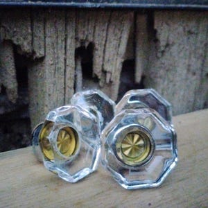 May include: A collection of clear, faceted glass cabinet knobs with gold-colored centers. The knobs are arranged on a wooden surface, with a weathered wooden background.