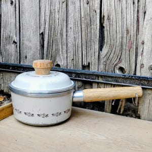 Puede incluir: Una cacerola vintage de color crema con tapa y asa de madera. La cacerola tiene un estampado floral alrededor de la base y un pomo de madera en la tapa. El asa es de madera clara y está sujeta con una banda metálica.