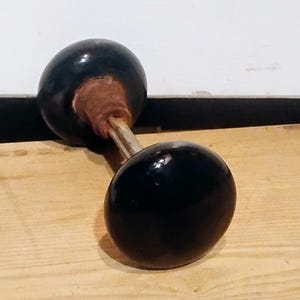 Puede incluir: Una mancuerna con dos pesas esféricas negras conectadas por una barra plateada. La sección central de la mancuerna es de color marrón rojizo. La mancuerna está sobre una superficie de madera clara.