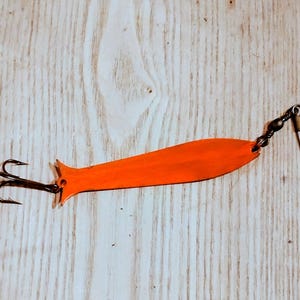 以下が含まれることがあります： オレンジ色の釣りルアー。トリプルフックとスイベルクラスプが付いています。ルアーは涙の形をしており、金属製です。フックは黒色で、スイベルは銀色です。このルアーは魚を引き寄せるように設計されています。