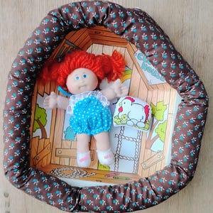 Può includere: Una bambola vintage Cabbage Patch Kids con capelli rossi di lana, occhi blu e un vestito blu a pois. La bambola è in un letto a fantasia marrone e bianco con un cuscino a tema pollo. Il letto ha un bordo marrone con un motivo floreale blu e bianco.