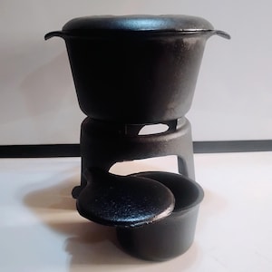 May include: A black cast iron fondue pot set. The set includes a pot with a lid, a stand, and a small pot with a lid. The fondue pot has a textured surface and is designed for cooking and serving. The set is made of durable cast iron.