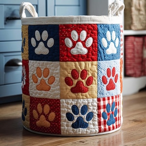 May include: A round, quilted storage basket with a patchwork design featuring paw prints. The squares are in various colors, including red, blue, and yellow, with white paw prints. The basket has white handles and a white rim.