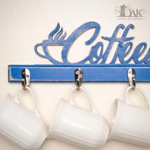 May include: A blue wooden wall-mounted rack with the word Coffee in cursive script and a steaming cup icon. The rack features three silver metal hooks, each holding a plain white ceramic mug. The blue paint has a distressed finish.