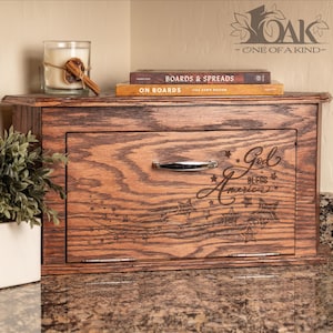 May include: A wooden bread box with a drawer, featuring the engraved text "God Bless America" and star designs. The box is stained a warm brown and has a silver handle. A candle and books are on top.