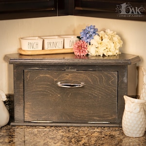 May include: A dark stained wooden bread box with a drawer and a decorative top. The top holds a wooden tray with three small white ceramic containers labeled "Pinch", "Sprinkle", and "Dash". A bouquet of flowers sits on the right.