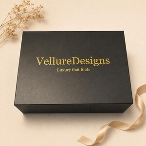 Könnte beinhalten: Eine rechteckige schwarze Geschenkbox mit dem goldenen Schriftzug VellureDesigns und dem Slogan Luxury that folds mittig auf dem Deckel. Die Box wird von einem hellbeigen Band und einem kleinen Zweig getrockneter Blumen in der Ecke begleitet.