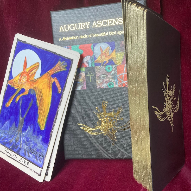 A photo of the card deck in colour . Thepack has gilded edges and the top card on display is of a golden winged hare with a blue background and a silver moon ,