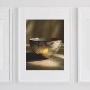 May include: A framed print of a Kintsugi bowl. The ceramic bowl is off-white with gold-colored veins and repairs. The bowl is centered in the frame, with a soft, warm light illuminating it.