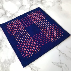 May include: A square piece of navy blue fabric with a geometric pattern stitched in pink and orange thread.