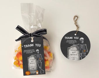 Puede incluir: Una bolsa de regalo con temática de Halloween llena de caramelos de maíz, atada con una cinta de lunares en blanco y negro y una etiqueta a juego. La etiqueta presenta un esqueleto, una lápida y las palabras "Gracias". También es visible una segunda etiqueta.