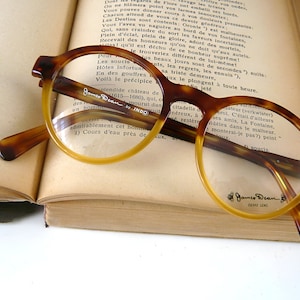 May include: A pair of tortoiseshell round spectacles with gold-rimmed lenses. The glasses are resting on an open book with text in French. The brand name "James Dean" is printed on the right lens.