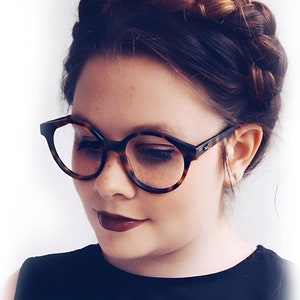 Könnte beinhalten: Eine Frau mit braunen Haaren, die zu einer geflochtenen Krone gestylt sind, trägt eine runde Brille aus Schildpatt und dunklen Lippenstift.