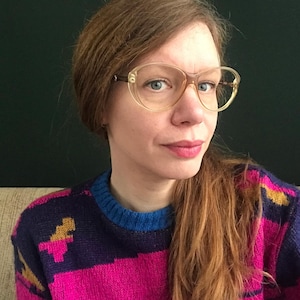 May include: A woman with long, light brown hair wearing a pink, purple, and blue striped sweater with a gold dinosaur design. She is wearing glasses with a gold frame.