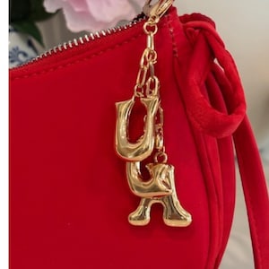 May include: A red handbag with a gold-colored chain and charm. The charm features the letters "U" and "A" in a gold, serif font. The bag has a zipper and a fabric tie detail.