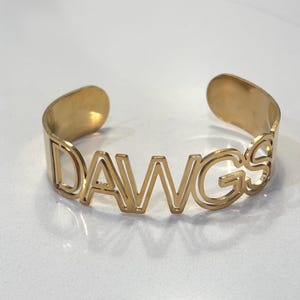 May include: A gold-toned metal cuff bracelet featuring the word DAWGS cut out in a bold, sans-serif font. The bracelet has an open-back design with rounded ends, showcasing a smooth, polished finish throughout the entire piece of jewelry.
