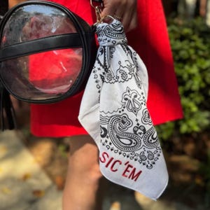 May include: A white bandana with a black paisley design and the red text "SIC 'EM" is attached to a black and clear round purse. The purse has a black strap and gold hardware. The background is a red dress.