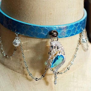 Op de afbeelding: Een blauwe choker ketting met een textuurpatroon, versierd met een zilveren hanger met een blauwe edelsteen en bungelende kettingen met kristallen accenten. De ketting wordt getoond op een mannequin.