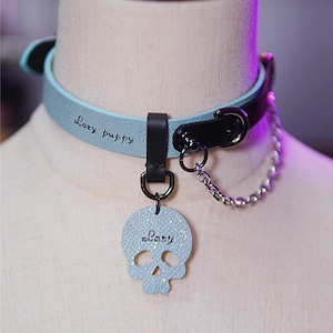 Op de afbeelding: Lichtblauwe en zwarte leren halsband met een schedelvormige bedel. De woorden "Lazy puppy" en "Lazy" staan op de halsband. Een zilveren ketting is aan de halsband bevestigd.