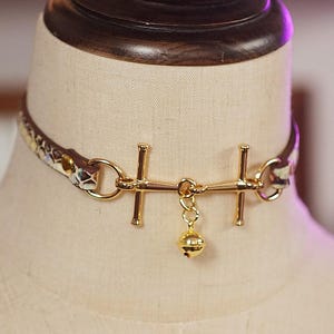 Op de afbeelding: Een goudkleurige choker-ketting met een kleine belhanger. De choker heeft een patroonband met gouden hardware en een decoratief middelpunt. De ketting wordt op een mannequin getoond.