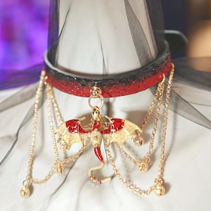 Op de afbeelding: Een rode en zwarte choker ketting met een gouden drakenhanger. De draak heeft rode en gouden vleugels en een rode staart. Gouden kettingen met kleine gouden accenten hangen van de choker.