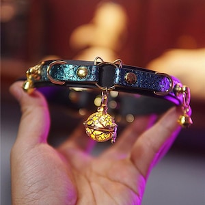 Op de afbeelding: Een kattenhalsband met een teal en zwart ontwerp, versierd met goudkleurige accenten en een decoratieve bel. De halsband heeft een kleine katvormige bedel en een gloeiende, sierlijke belhanger. De halsband wordt in een hand gehouden.