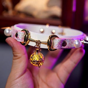 Op de afbeelding: Een witte halsband met goudkleurige hardware, een decoratieve hanger en parelaccenten. De halsband heeft een patroon en is versierd met een goudkleurige, bolvormige hanger met een ingewikkeld, verlicht ontwerp.