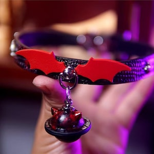 Op de afbeelding: Zwarte dierenhalsband met rode vleermuisvleugelaccenten en een bijpassende bedel. De halsband heeft een textuurpatroon en zilverkleurige hardware. De bedel heeft een donkerrood en zwart ontwerp.