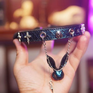 Op de afbeelding: Een donkerblauwe choker-stijl kraag met zilverkleurige kruis- en schedelaccenten. Een bungelende bedel met konijnenoren en een druppelvormige kraal hangt in het midden. De kraag wordt in een hand gehouden.