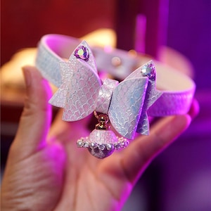 Op de afbeelding: Een lichtblauwe en witte dierenhalsband met een strik en een bungelende bedel. De halsband heeft een textuur, schubachtig patroon. De strik is versierd met een sprankelende edelsteen. De bedel is een kleine, decoratieve bol.