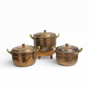 May include: Three antique copper pots with lids and brass handles. The pots vary in size, with a larger one on a small wooden stand. The copper has a rich, aged patina, suggesting vintage cookware.