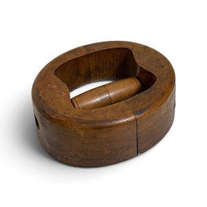 May include: A dark brown wooden hat block with an oval shape and a central horizontal handle. The surface shows natural wood grain and signs of wear, featuring a hollowed-out center and a smooth, rounded exterior finish for shaping headwear.