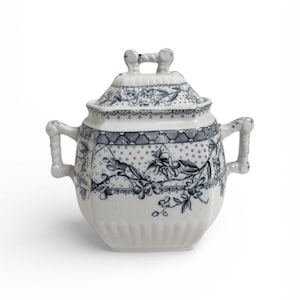 May include: A white and blue ceramic sugar bowl with a lid. The bowl has two handles and a floral design. The lid has a handle and a matching floral pattern. The sugar bowl is a vintage style and is likely made of porcelain.