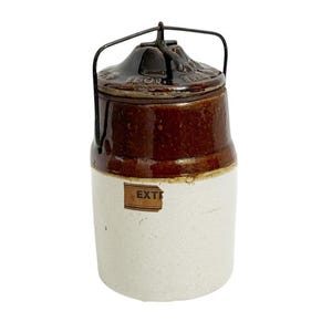 May include: A cylindrical stoneware crock with a two-tone finish, featuring a dark brown glazed upper section and a cream-colored lower base. It includes a matching brown lid with a metal wire bail handle and a partial label reading EXT.
