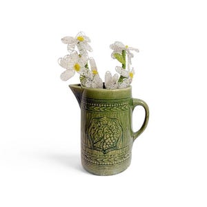 May include: A green ceramic pitcher with a textured surface and a central embossed emblem, containing a bouquet of white and yellow wire-beaded flowers with green stems. The pitcher features a handle and a pouring spout on the upper rim.