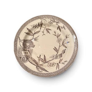 May include: A round ceramic plate with a cream-colored base and dark brown transferware patterns. The design features a bird perched on a branch, floral elements, bamboo-like leaves, and rectangular motifs. The rim has a decorative lattice border.