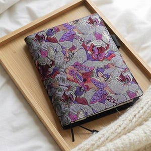 May include: A journal with a floral pattern in purple, red, and grey, sits on a light wooden tray. The journal is on a white bed with a cream-coloured knitted blanket.
