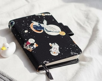 May include: A black notebook with a space-themed design featuring astronauts and planets. The notebook has a black leather cover and a black ribbon bookmark. A small figurine and a white duck figurine are next to the notebook.
