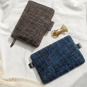 May include: Two rectangular fabric cases, one brown and one blue, with black leather tabs. A gold-colored decorative hand-shaped object is also present. The items are arranged on a white surface.