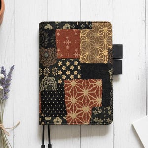 May include: A rectangular fabric notebook cover featuring a patchwork design of black, rust-orange, and gold-toned fabric panels. The patterns include floral motifs, geometric star shapes, and small triangular prints. Two black pen loops extend from the side.