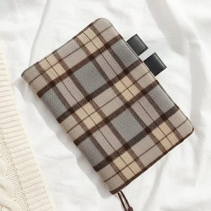 Op de afbeelding: Een rechthoekige etui met een ruitpatroon in grijstinten, beige en bruin. Het heeft een leren lipsluiting en een trekkoord. De etui ligt op een witte ondergrond, naast een crèmekleurige gebreide trui.