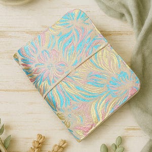 May include: A beige notebook with a floral design in pink, blue, and gold. The notebook is closed with an elastic band. The image is set on a light wood surface with decorative plants and a green cloth.