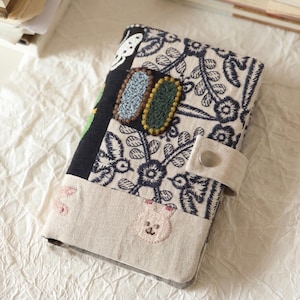 Op de afbeelding: Een rechthoekige stoffen notitieboekhoes met een patchworkontwerp, voorzien van een zwart-wit bloemenpatroon, een beige gedeelte met geborduurde roze konijnengezichtjes en twee ovale, getextureerde patches in blauw en donkergroen, vastgezet met een drukknoop.